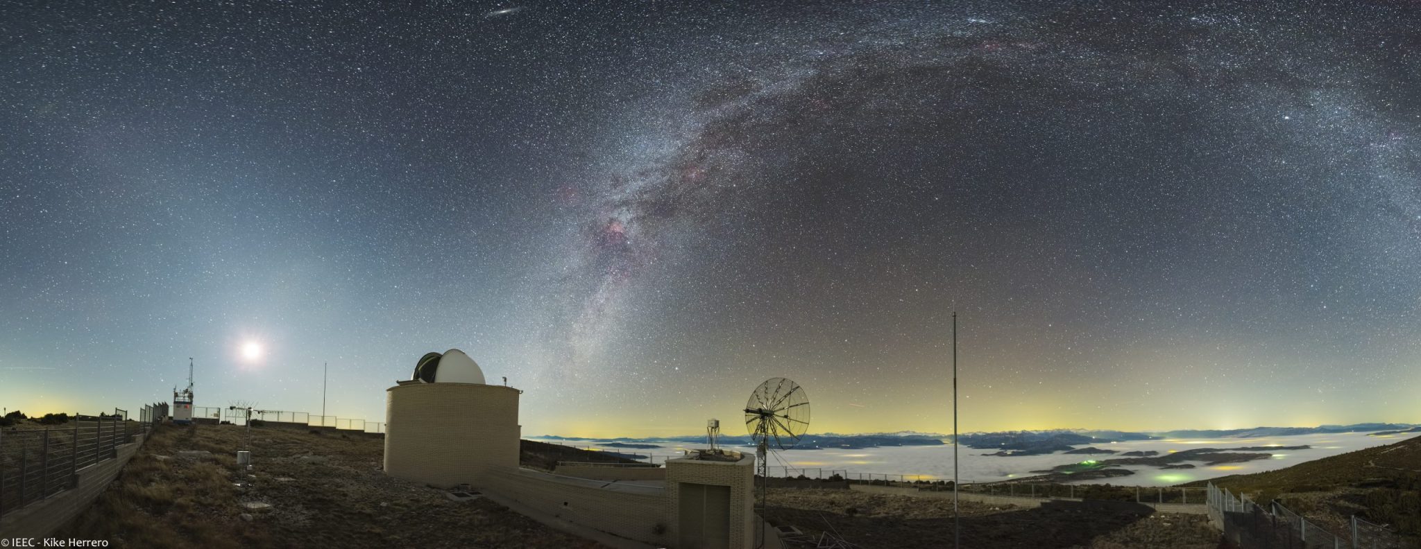 Panoramic night photograph of Montsec Observatory, with its dome and radio antenna beneath the Milky Way above the surrounding mountains.